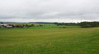 Blick über eine wellige Landschaft mit Wiesen und Viehweiden. Links im Hintergrund, hinter Siedlungshäusern, sind auch Äcker erkennbar. Rechts befindet sich ein Waldsaum, hier stehen Windräder.
