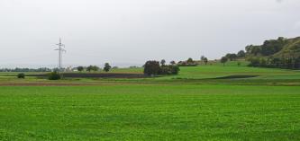 Das Bild zeigt flache grüne und braune Äcker im Vordergrund. Dahinter folgt eine stufige, nach rechts ansteigende Geländeerhöhung mit schwarzen Ackerböden und grünen Wiesen. Ganz rechts ist noch ein bewaldeter Berg angeschnitten.