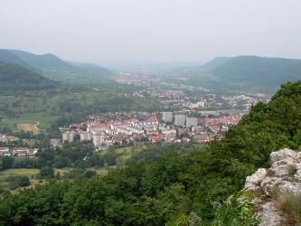 Von erhöhtem Standpunkt aus hat man - über Felsen und Baumwipfel - einen weiten Blick auf ein Tal und mehrere, fast zusammenhängende Ortschaften, die bis zum Horizont reichen. Links und rechts grenzen bewaldete Höhenzüge an.