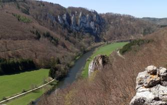 Von erhöhtem Standpunkt aus sieht man auf den Bogen eines Flusses, der rechts hinter Wald verschwindet. Links des Flusses folgen bewaldete Hänge seinem Lauf. In der Hangmitte türmen sich steile, grauweiße Felsnadeln auf.