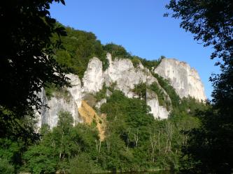 Oberhalb eines gerade noch erkennbaren Flusses und inmitten dichter Bewaldung erheben sich mehrere schroffe, steile, weißlich graue Felstürme. In der Bildmitte links ist zudem eine gelblich gefärbte Schutthalde zu sehen.