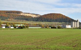 Hinter einer ebenen, begrünten Ackerfläche und Industriebauten steigen grüne und braune Hänge sowie bewaldete Höhenzüge in Herbstfarben auf. In der Mitte ist über den Baumspitzen ein offener Steinbruch erkennbar.