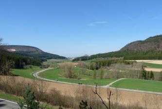 Das Bild zeigt eine wellige, aus Äckern und Grünland bestehende Ebene, die von Bäumen und einer Straße durchzogen ist. Im Hintergrund erheben sich links und rechts bewaldete Steilhänge. Links ist zudem ein Steinbruch erkennbar.