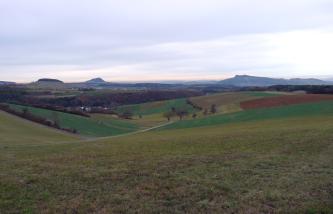 Das Bild zeigt eine weite, von links nach rechts erst absinkende und dann wieder ansteigende Acker- und Grünlandschaft. Im Hintergrund sind ein bewaldeter Hügel sowie zwei Vulkanberge erkennbar.