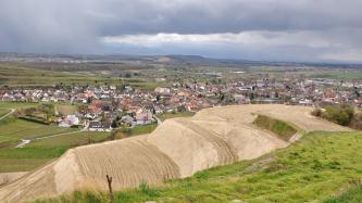 Im Vordergrund des Bildes sind stufige Lössterrassen zu sehen, die nach rechts ansteigen, sowie Grünland. Zum Hintergrund hin breitet sich unter einem Gewitterhimmel eine Landschaft mit Feldern, Hügeln, Bergen und großflächiger Besiedelung aus.
