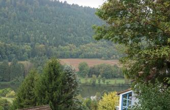 Entlang des hier ruhig fließenden Neckars sind von Bäumen bestandene Ufer und angrenzende Ackerflächen sichtbar. Dahinter steigt steil ein bewaldeter Berg auf.