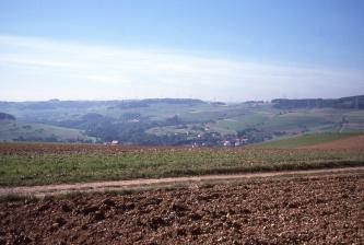 Das Bild zeigt mehrere im Vordergrund liegende Äcker sowie im Hintergrund ein tiefes, welliges Tal mit Acker-, Grün- und Waldflächen.