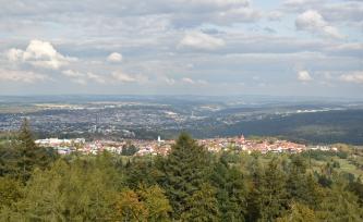 Blick aus großer Höhe über Baumwipfel und bewaldete Bergrücken. Im Hintergrund breitet sich eine größere Stadt aus. Im Mittelgrund, auf einem Bergsattel, liegt eine Ortschaft mit Kirchtürmen.
