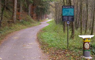 Am Eingang zum Geologischen Pfad im Hölltal südwestlich von Schwäbisch Gmünd Blick auf einen geteerten, kurvigen Weg, der in einen herbstlichen Wald führt. Rechts steht ein Hinweisschild mit der Überschrift „Geologischer Pfad“; weiter vorn zeigt sich eine bemalte Figur.