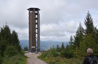 Blick auf einen hohen, sechseckigen Aussichtsturm am Rand einer Bergkuppe. Der Turm hat teils Wände aus Schindeln, teils ist er offen, so dass der Treppenaufgang sichtbar ist. Am Fuß des Turmes verläuft ein Wanderweg. Links und rechts stehen Nadelbäume.