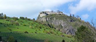 Hinter einem nach rechts abfallenden grünen Hügel, der teils mit Bäumen bestanden, teils mit Rebstöcken bepflanzt ist, erhebt sich rechts ein länglicher Felsenberg. Auf der flachen Kuppe des Berges erstreckt sich eine Burgruine.