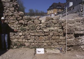 Blick auf rötlich graue, ältere Mauerreste am Rand einer Baugrube. Die Mauersteine haben unterschiedliche Formen und Farben. Links steht ein Mann mit Helm und weist auf ein Schild sowie eine Messlatte hin.