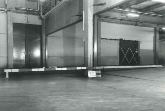 Ältere Schwarzweißaufnahme vom Inneren einer Fabrikhalle. Der Boden der Halle zeigt in der Mitte Sprünge und Risse. Eine quer aufgelegte Markierungsschranke weist rechts eine Aufwölbung des Bodens von 23 Zentimetern gegenüber links aus.