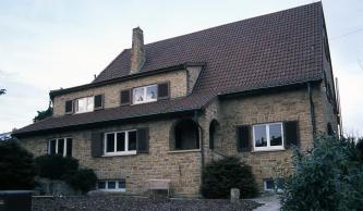 Das Foto zeigt die Langseite eines Wohnhauses mit Dachgaube und spitzem Dach. Das Mauerwerk des Hauses besteht aus gelblich braunen Steinen.
