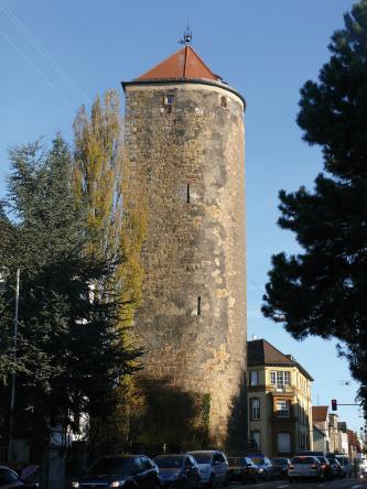 Das Bild zeigt einen hohen Steinturm am Ende einer Häuserzeile. Der Turm ist gelblich bis graubraun, hat schmale Schlitze und kleine Fenster sowie ein dreieckiges Ziegeldach.