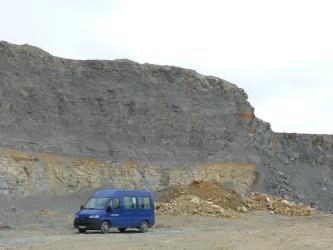 Blick auf eine hohe, nach rechts abfallende Abbauwand in einem Steinbruch. Das untere Viertel besteht aus gelblich-beigen gebankten Gesteinen, darüber befindet sich dunkelgraues bis blaugraues Gestein. Vor der Wand steht ein blauer Van.