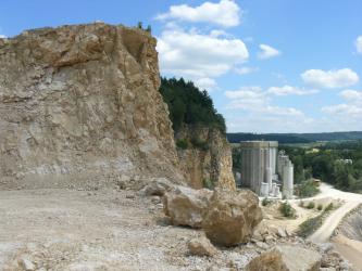 Gesteinsgewinnung findet im Steinbruch Eigeltingen durch Reißen und Baggern statt. Blick von schräg oben in einen Steinbruch: Im Vordergrund links befindet sich eine Abbauwand aus hellgelbem Gestein, im Hintergrund rechts sind Industrieanlagen und dahinter Landschaft zu erkennen.