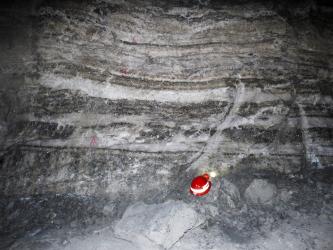 Blick auf eine Steinsalzwand in einem Bergwerk, hellgrau, weiß und dunkelgrau gestreift. Unten dient ein rot-weißer Helm als Größenvergleich.