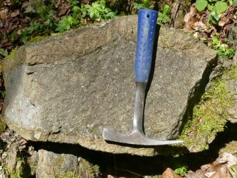 Das Foto zeigt einen angewitterten Gesteinsblock. Das Gestein ist mittel- bis dunkelgrau, teilweise von Moos bewachsen. Auf dem Block liegt ein Hammer mit blauem Griff.