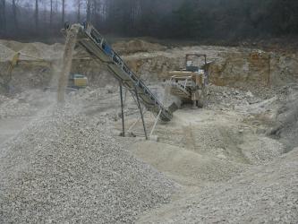 Kies und Sand werden maschinell abgegraben, der Kalkstein wird auch gesprengt. In der Bildmitte ist der maschinelle Abbau von Kiesen und Sanden durch einen Bagger zu sehen. Das Material wird auf ein Förderband gebracht und im Vordergrund des Bildes zu einem Hügel aufgeschüttet.