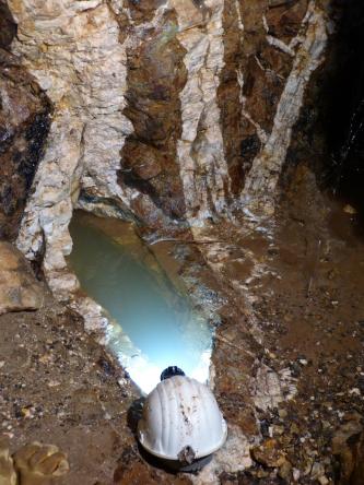 Blick auf einen dunkelbraunen Grubengang in einem Bergwerk. Von einem Helm ausgehend, der unten abgelegt wurde, und einer angrenzenden ovalen Wasserfläche, breiten sich wie Federn weißliche und braune Adern nach oben aus.