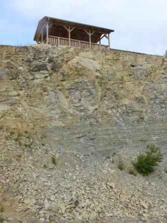 Blick von seitlich unten auf eine Gesteinswand, welche den Übergang zwischen bankigem, hellbeigem Gestein (oben) und massigem, grauem Gestein (unten) zeigt. Über der Wand steht eine offene Hütte aus Holz.