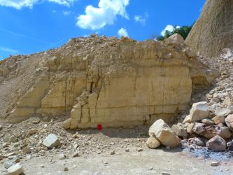 Blick auf eine Steinbruchwand aus dickbankigen (bis 1 Meter) hellen Kalksteinen. Rechts und links vor der Abbauwand liegen Gerölle. Der Himmel ist intensiv blau.
