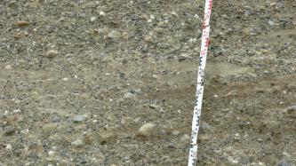 Das Bild zeigt gröbere und feinere Kiese, die in hellbraunem Bodenmaterial eingebacken sind. Ein Maßstab rechts zeigt die Größenverhältnisse an.