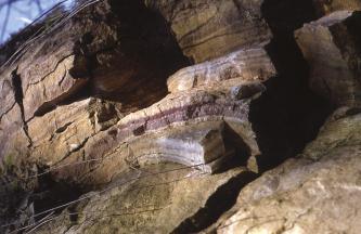 Nahaufnahme einer abgestuften, mit Nischen und Stufen versehenen Gesteinsschicht. Im Vordergrund verläuft ein bogenförmiger, violetter, streifiger Aufsatz.