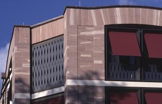Blick auf den oberen Teil einer Gebäudefassade aus rötlichen Mauersteinen mit hellgrauen Streifen. Rechts sind mehrere Fenster mit Sonnenschutzblenden angeschnitten; unterbrochen von einem horizontal verlaufenden weißen Sims.