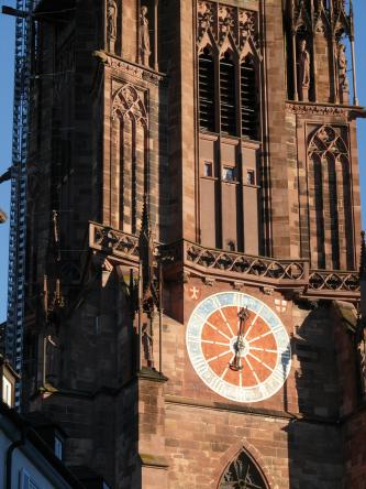 Teilansicht des Freiburger Münsterturms aus rotbraunem Gestein mit Ornamenten und Spitzbogenfenstern. Unten rechts ist das Zifferblatt der Münsteruhr zu sehen.