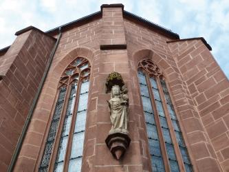 Aufwärts blickend sieht man hier die rötliche Steinfassade einer Kirche mit hohen Spitzbogenfenstern und Stützpfeilern. Zwischen den Fenstern, etwas unterhalb der Bildmitte, ist eine Frauenfigur angebracht.