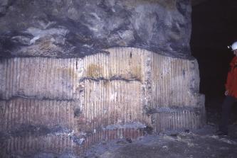 Blick auf eine teilweise offenliegende Gesteinswand innerhalb eines Bergwerkes. Der offene Teil weist zahlreiche senkrecht verlaufende Furchen auf; die Mitte ist zudem von einer Lampe beleuchtet, die ein Bergmann rechts auf seinem Helm trägt.