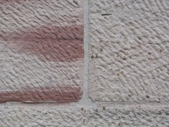 Nahaufnahme von rötlich grauen bis weißlichen Mauersteinen mit geriffelter Oberfläche. Links im Bild sind größere rote Stellen erkennbar.