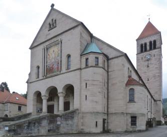 Blick auf Stirnfront und rechte Seite einer Kirche mit Säuleneingang und angebauten, gerundeten Türmchen. Die Kirche ist aus rötlich grauem Stein gebaut. Hinten rechts erhebt sich ein viereckiger Glockenturm mit rotem Spitzdach.
