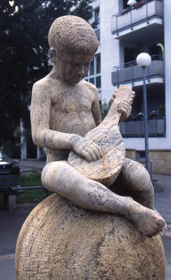 Die Aufnahme zeigt eine Steinfigur, sitzend auf einer Steinkugel. Figur und Kugel sind aus gelblichem bis grauem Material; die Figur ist einem Jungen nachgebildet, der ein Zupfinstrument in den Händen hält.