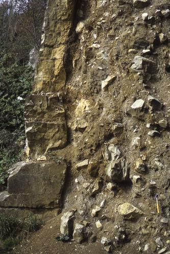 Blick auf eine senkrecht aufragende, braune Erdmauer, an die sich links Felsblöcke anlehnen. Rechts sind Gesteinsbrocken in das Erdreich eingebettet.