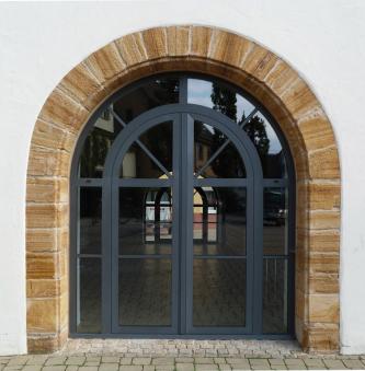 Blick auf ein modernes, aus Glas und Metall gefertigtes, oben abgerundetes Eingangsportal eines Gebäudes. Das Portal umgibt ein schmaler Kranz aus rötlichem bis grauem Gestein. Der sichtbare Rest der Fassade ist weiß.