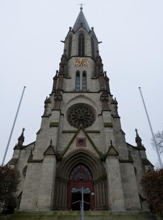 Blick auf den Eingangsbereich einer Kirche mit sich darüber erhebendem Turm aus grauem Gestein. Der Kirchturm wird von mehreren seitlichen Pfeilern gestützt und dem neugotischen Baustil zugeordnet.