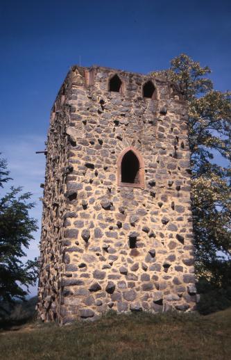 Das Bild zeigt die Front eines viereckigen Turms, welcher auf einer kleinen Erhöhung steht. Der Turm ist aus rötlich bis gelblich grauen Gesteinen gebaut und hat ein großes Fenster in der Mitte und oben noch kleinere Fenster.