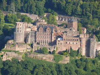 Blick von erhöhtem Standort auf die rötlichen Mauern von Schloss Heidelberg. Die Türme links und rechts außen sowie ein Anbau sind für Sanierungsarbeiten eingerüstet.
