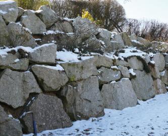 Blick auf mehrere roh behauene, graue Steinblöcke, die zu einer getreppten Mauer aufgeschichtet sind. Die Rohblöcke sind an den Oberseiten teils von Schnee bedeckt.