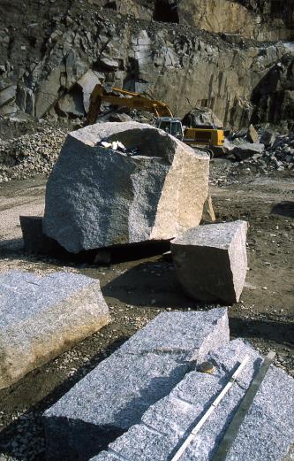 Blick auf mehrere teils flache, teils würfelförmige graue Steinblöcke in einem Steinbruch. Im Hintergrund arbeitet ein Bagger vor einer Gesteinswand.