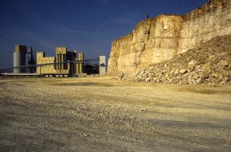 Blick auf eine Steinbruchwand aus gelblich braunem Gestein sowie einem Abraumhaufen davor (rechts im Bild). Im Hintergrund links sind Förder- und Verarbeitungsanlagen zu sehen.