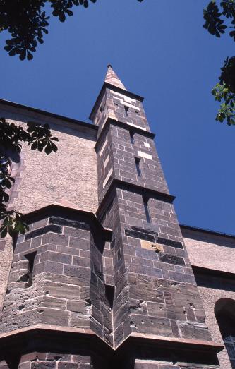 Teilansicht einer Kirchenfassade mit angesetztem, schmalem Turm aus dunkelgrauem Mauerwerk. Der Turm ist mehrfach unterteilt und hat schmale Fensterschlitze.