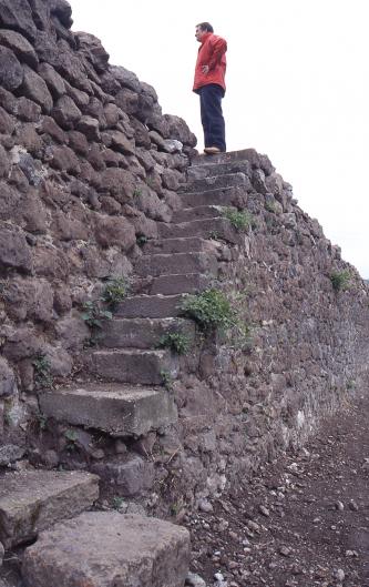 Das Foto zeigt in Schrägansicht eine hohe Steinmauer, mit Fluchtrichtung rechts. Eine angebaute Steintreppe führt auf die Mauerkrone. Auf der obersten Stufe steht ein Mann mit roter Jacke.