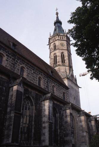 Teilansicht einer Kirche mit Haupt- und Seitenschiff aus dunkelgrauem Gestein sowie Turm (rechts im Bild) aus gelblich grauen Steinen.