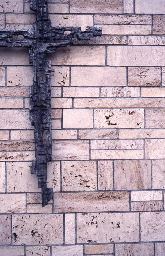 Nahaufnahme einer Wandverkleidung mit rötlich grauen Steinplatten, ähnlich Kacheln, in unregelmäßigem Muster. Links hängt ein stilisiertes, dunkelgraues Metallkreuz an der Wand.