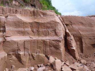 Das Bild zeigt zwei versetzt stehende Steinbruchwände. Auf dem rötlich braunen Gestein sind links mehrere senkrecht verlaufende Furchen zu erkennen. Unten rechts liegt ein Hammer auf einem losen Block.