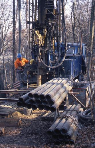 Das Foto zeigt ein großes Bohrgerät mit Arbeiter. Im Vordergrund, auf einem Gestell und am Boden, liegen mehrere dünne Rohre. Im Hintergrund stehen hohe, dünne Bäume.